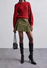 Personne portant un pull en tricot rouge, une mini-jupe boutonnée vert olive, des bottes à talons noires montant jusqu'aux genoux, et tenant un petit sac à main noir.