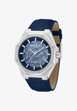 Kenneth Colen automaattikello, jossa hopeinen kahdeksankulmainen kotelo, sininen skeleton-taulu, hopeiset tuntimerkinnät ja teksturoidulla sinisellä kangasrannekkeella.