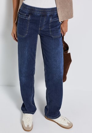 Mørkeblå denim bukser med elastisk talje, der har forlommer og lige ben. Båret med hvide sneakers og en brun taske.