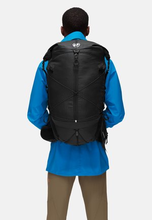 Black backpack with textured fabric, adjustable straps, and a bungee cord design on the back. Paired with a blue long-sleeve shirt.