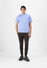 Helle blaue Baumwoll-T-Shirt mit geripptem Hals, braune eng anliegende Jogginghosen und dunkle Sneakers mit strukturiertem Sohlen, vor einem weißen Hintergrund stehend.