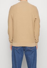 Beige strukturierter Pullover mit Rippmuster, Rundhalsausschnitt und eng anliegenden Bündchen, kombiniert mit lässigen blauen Jeans.