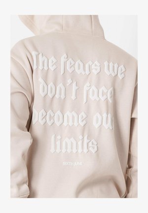 Lys beige hoodie med en tekstureret hvid grafik på ryggen, der har fed, stiliseret tekst. Inkluderer en hætte og en blød stofstruktur.
