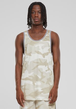 Jeune homme aux dreadlocks mi-longs portant un maillot de sport sans manches camouflage beige et blanc avec un short assorti sur un fond uni.