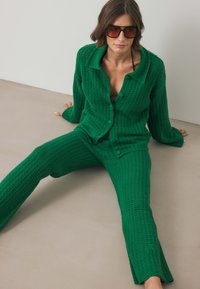 Zaļš adīts komplekts, kas sastāv no pogājama, ilgiem piedurkņu džemperiem un atbilstošiem biksēm ar teksturētu apdari. Modelei ir uzliktas pārmērīga izmēra saulesbrilles.