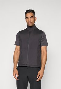 Man in een zwart mouwloos zip-up vest over een kortekleurig donkergrijs shirt, staand tegen een effen lichte achtergrond.