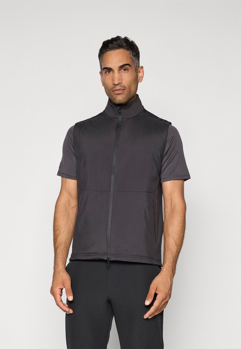Man in een zwart mouwloos zip-up vest over een kortekleurig donkergrijs shirt, staand tegen een effen lichte achtergrond.