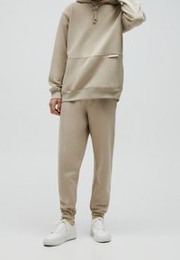 Sweat à capuche beige avec une poche kangourou, une fermeture à cordon de serrage et un pantalon de survêtement assorti à coupe fuselée. Associé à des baskets blanches. Texture lisse.