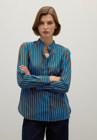 Woman with short hair wearing a blue and brown vertical striped button-up shirt, gold chain necklace, arms crossed, standing against a plain wall.