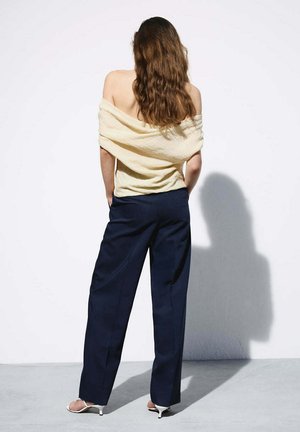 Femme aux longs cheveux bruns, debout avec les mains dans les poches, portant un haut crème à épaules dénudées, un pantalon large bleu marine et des sandales blanches à talons.