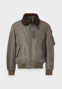 Brun krave bomberjakke i olivengrøn, med lynlåslukning foran, sidelommer og ribbede manchetter og kant. Materialet virker glat med en mat finish.