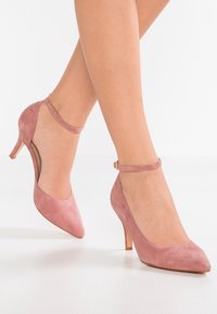 Rosa mockaskinnshöga klackar med spetsig tå och ankelrem. Har en smal klack och utklippda detaljer på sidorna. Slät textur.