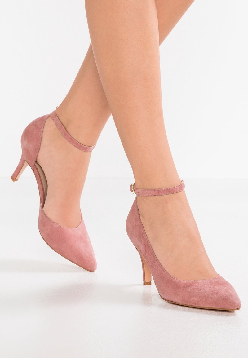 Rosa mockaskinnshöga klackar med spetsig tå och ankelrem. Har en smal klack och utklippda detaljer på sidorna. Slät textur.