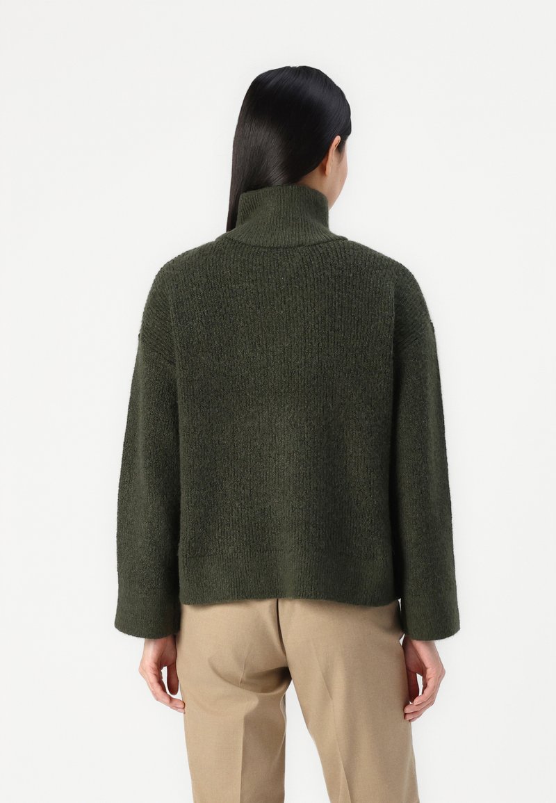 Grüner, grob gestrickter Pullover mit hohem Kragen und weiten Ärmeln. Weiche Textur, cropped Länge und gerippte Details am Kragen und an den Bündchen.