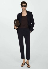 Blazer alla marinara su una canotta a coste marrone, abbinato a pantaloni slim-fit neri e infradito neri, mentre tiene una pochette marrone.