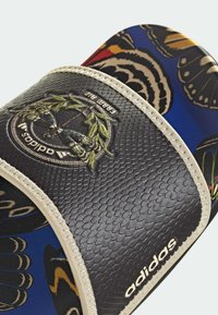 Glissière synthétique noire à texture, avec un patch logo, entourée d'un motif de papillons colorés sur un fond bleu et des accents beiges.