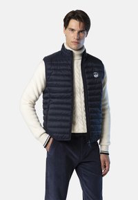 Mann in einer marineblauen Steppweste über einem weißen, grob gestrickten Rollkragenpullover und marineblauen Cordhosen, stehend mit einer Hand in der Tasche.