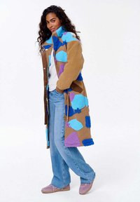 Kobieta ubrana w długi brązowy polarowy płaszcz z niebieskimi i fioletowymi łatami, białą bluzkę, jasnoniebieskie dżinsy i różowe trampki, stoi z dłońmi w kieszeniach.