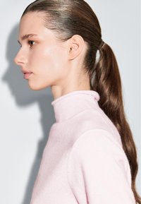 Maglione a collo alto rosa chiaro con una consistenza morbida. Il modello ha capelli lunghi e lisci di colore castano, pettinati all'indietro. Sfondo neutro.
