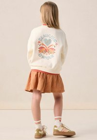 Sweatshirt crème avec des graphismes de flamant rose colorés et de cœurs, associé à une jupe plissée orange et des baskets vert clair avec des accents blancs.
