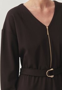 Zwarte jumpsuit met een V-hals en gouden rits, met een fitted taille geaccentueerd door een bijpassende riem en metalen gesp detail.