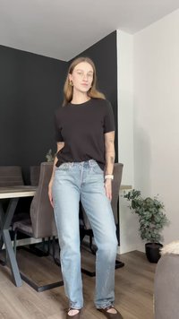 Črna majica s kratkimi rokavi, svetlo modri naravnost krojeni kavbojki z rahlim leskom, v kombinaciji z rjavimi balerinkami. Minimalni dodatki, sodoben ambient.