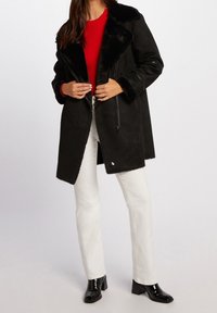 Sort faux fur-foret dyne med en bred krave, lynlås lukning og lange ærmer, båret over en rød ribbet sweater og hvide bukser.