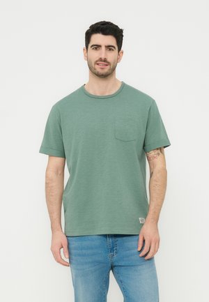 Homme aux cheveux courts et foncés avec une barbe, portant un t-shirt vert à manches courtes et un jean bleu, debout devant un fond blanc uni.
