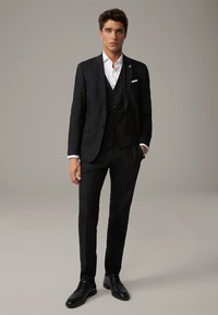 Uomo in abito nero a tre pezzi con camicia bianca, scarpe eleganti nere, in piedi su uno sfondo grigio uniforme.