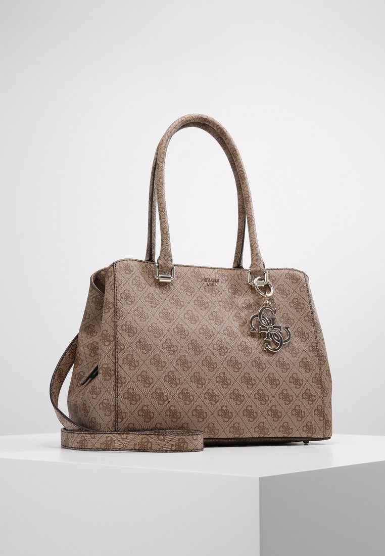 Sac à main beige à motif avec double poignées, bandoulière amovible, et breloque logo en métal suspendue à une poignée, posé sur une surface blanche.