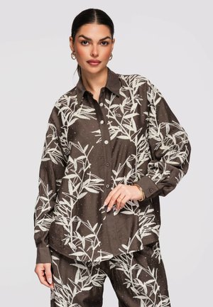 Vrouw draagt een loszittend bruin hemd- en broekenset met witte bladpatronen, staand tegen een effen lichte achtergrond.