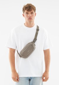 Borsa a tracolla grigia in tessuto con chiusura zip, tracolla regolabile e dettaglio con logo, indossata sul petto sopra una maglietta bianca.