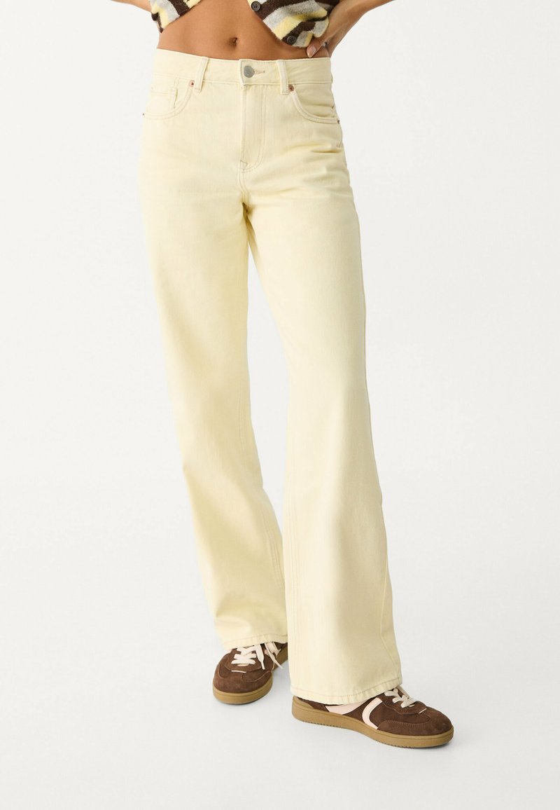 Jeans évasés de couleur jaune clair en coton avec une texture lisse. Dispose d'un design classique à cinq poches et d'une fermeture à bouton en métal.