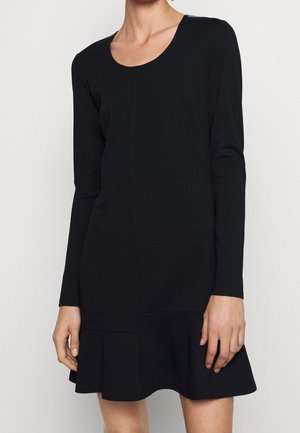 Vrouw die een effen zwarte jurk met lange mouwen draagt, met een ronde halslijn en een uitlopende zoom, staand tegen een witte achtergrond.