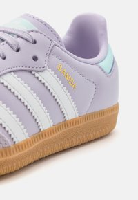 Baskets en cuir violet clair avec trois bandes blanches, texte jaune "SAMBA" et une semelle en caoutchouc colorée marron clair avec des motifs texturés.