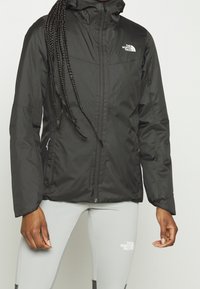 Černá voděodolná bunda s předním zipem, kapucí a logem. Má dlouhé rukávy a lehkou, hladkou texturu.