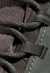 Primo piano di una scarpa nera con tessuto texture, lacci intrecciati e dettagli lisci, con una linguetta a fantasia e cuciture decorate.