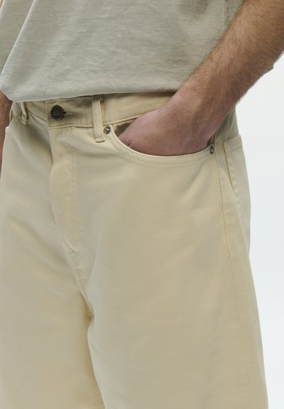 Osoba ubrana w jasnobeżowe spodnie z ręką wsuniętą do przedniej kieszeni, w połączeniu z jasnobeżową bluzką z krótkim rękawem.