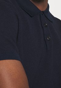 Mörkblå pikétröja med strukturerad stickning, korta ärmar och en treknapplåsning. Har ribbade mudd och krage.