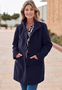 Marineblauer Mantel aus strukturiertem Stoff, mit Kragen, Knopfverschluss und seitlichen Taschen. Getragen über einem gemusterten Oberteil und Jeans.