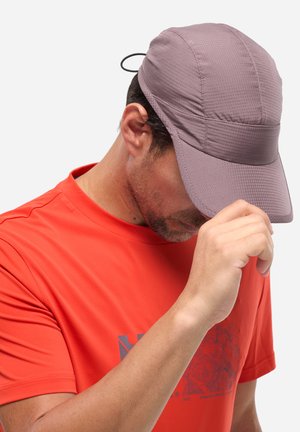 Homme portant une casquette mauve texturée et un t-shirt orange vif, ajustant la visière de la casquette de la main droite, tête légèrement inclinée vers le bas.