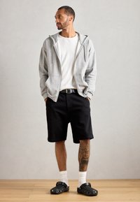 Siva zip-up dukserica, bijela majica, crne kratke hlače, crne cipele, bijele čarape. Značajne tetovaže na lijevoj nozi. Jednostavna, atletskog stila.