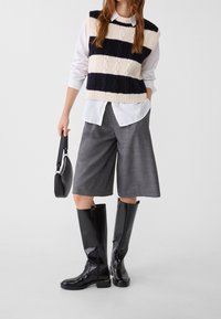 Gilet tricoté crème et rayures bleu marine sur une chemise blanche, shorts gris ajustés et bottes noires montantes, avec un sac à main noir.
