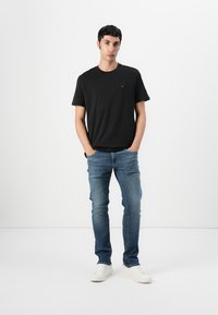 Black cotton t-shirt with a crew neck, paired with light blue denim jeans and white sneakers, standing against a plain background.