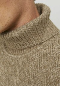 Maglione beige lavorato a maglia con un colletto alto a coste, caratterizzato da un motivo a spina di pesce testurizzato e un tessuto morbido e caldo.