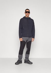 Svart zip-up jakke med hette, grå og svarte panelbukser, og svarte robuste sko. Modellen har på seg store mørke solbriller. En enkel hvit bakgrunn.