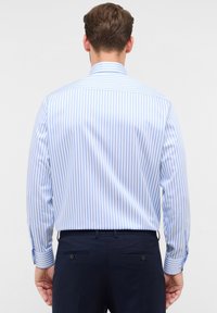 Uomo visto da dietro che indossa una camicia a righe verticale blu chiaro e bianca e pantaloni blu navy su uno sfondo semplice.