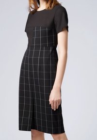 Černé šaty s krátkými rukávy; upnutá sukně s jednobarevným vrškem a kostičkovaným vzorem níže, který obsahuje bílé mřížkování a přední rozparek.