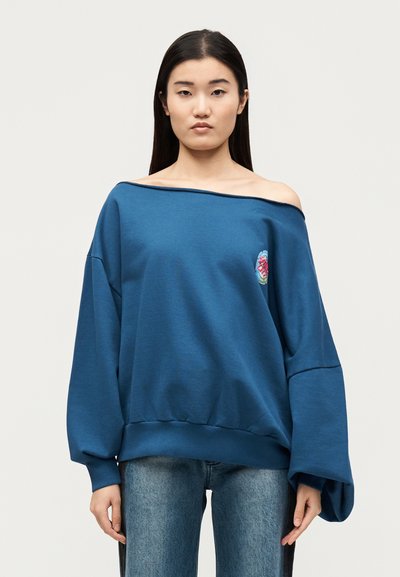 Γυναίκα με μπλε φαρδιά μπλούζα off-shoulder με μανίκια με όγκο και ανοιχτό μπλε τζιν μπροστά σε μονόχρωμο φόντο.