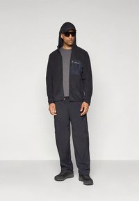 Črna flis jakna z zadrgo na prsnem žepu čez sivo majico, kombinirana s temnimi cargo hlačami in črnimi čevlji, z kapuco in sončnimi očali.